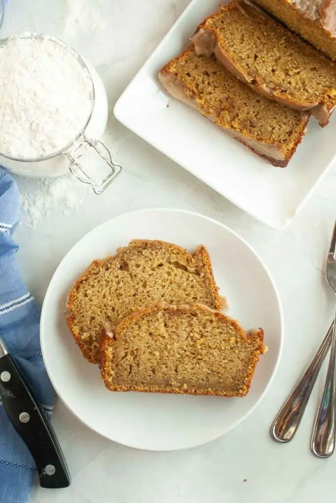 Two slices of banana bread on a white plate, with more sliced banana bread on a tray nearby, a jar of flour, utensils, and a blue cloth on a light surface.