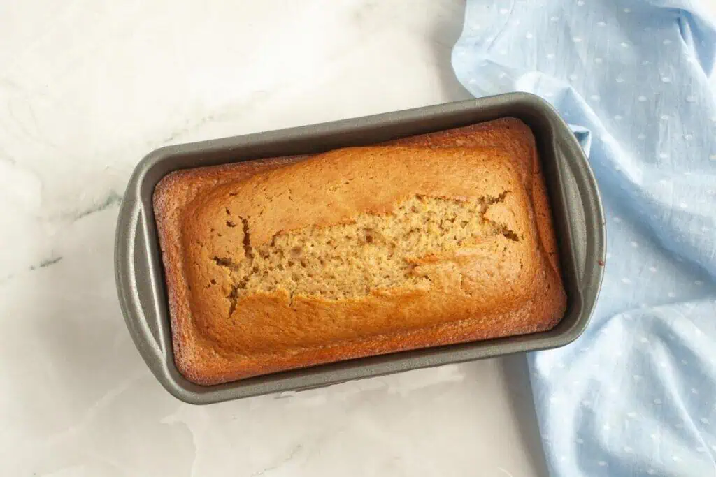 A loaf of banana bread in a rectangular metal baking pan sits on a white surface next to a light blue cloth with white polka dots.