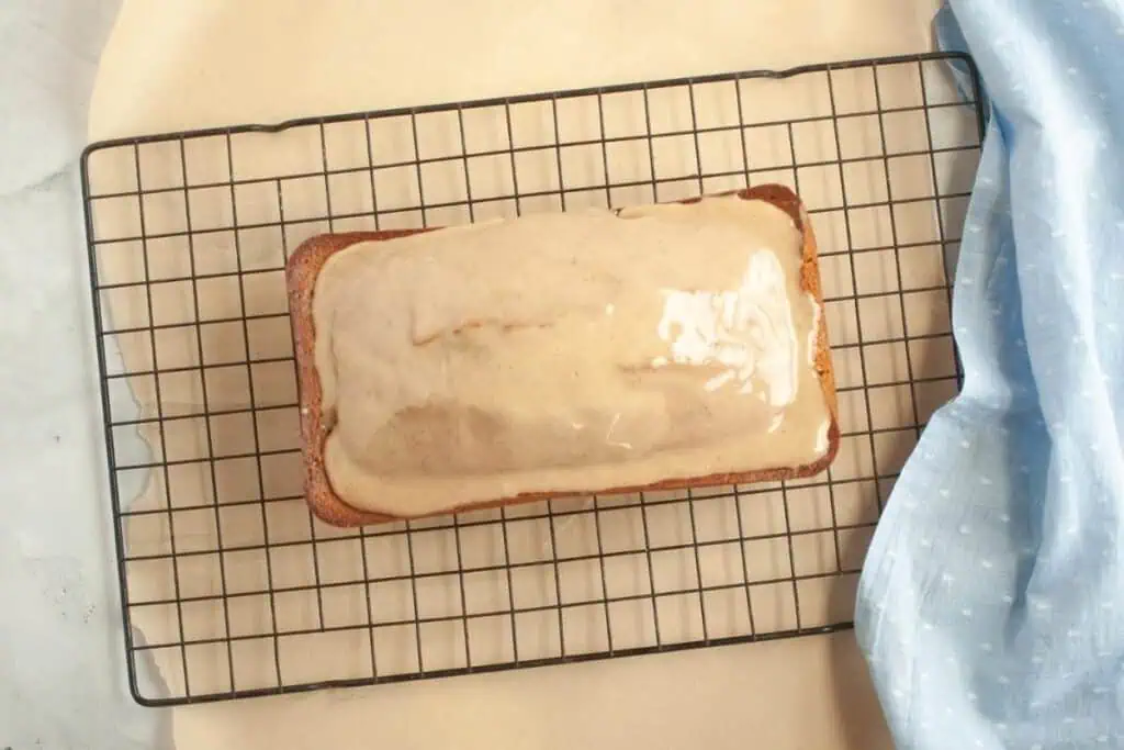 A loaf cake with light glaze sits on a cooling rack next to a blue cloth with white polka dots.