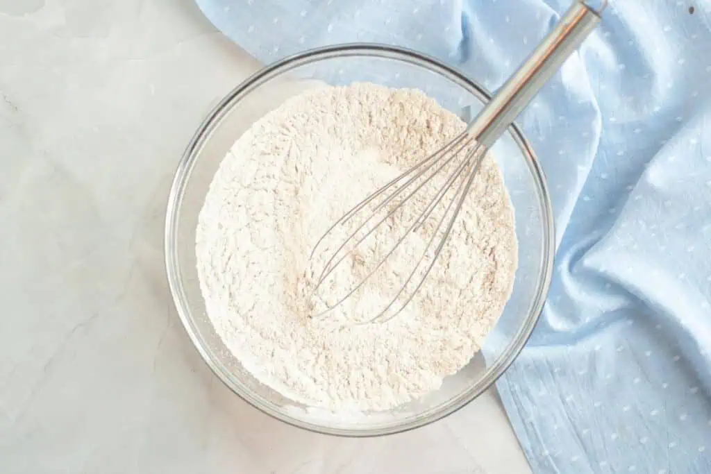 A glass bowl filled with flour mixture and a metal whisk sits on a light surface next to a blue dotted cloth.