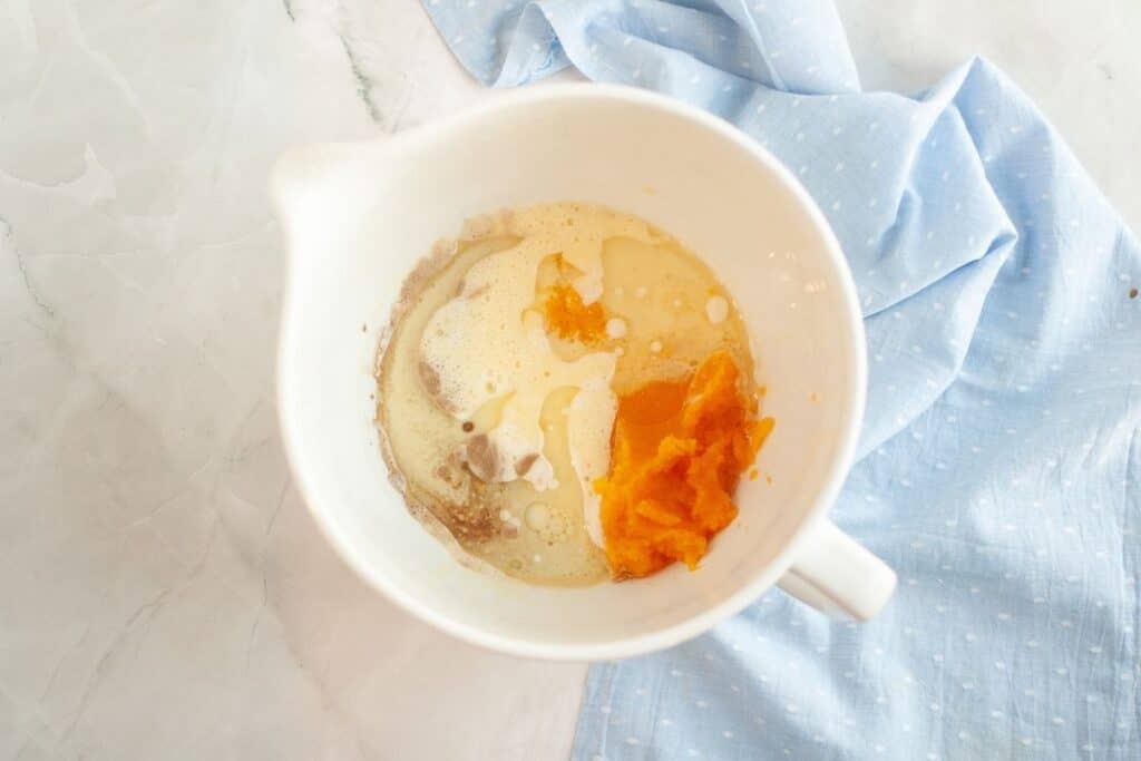 A white mixing bowl containing pumpkin puree, milk, and other baking ingredients sits on a marble surface next to a light blue cloth.