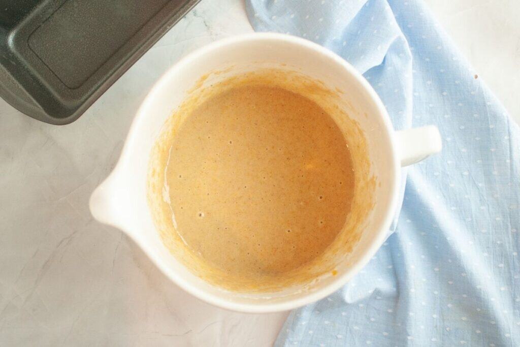 A white mixing bowl filled with light brown batter sits on a light surface next to a blue cloth and part of a metal pan.