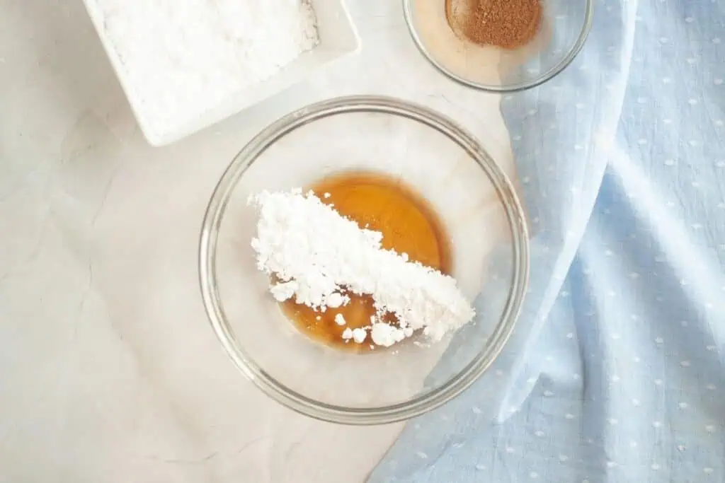 A glass bowl with powdered sugar and a brown liquid, next to a bowl of brown powder and a blue polka-dot cloth on a light surface.