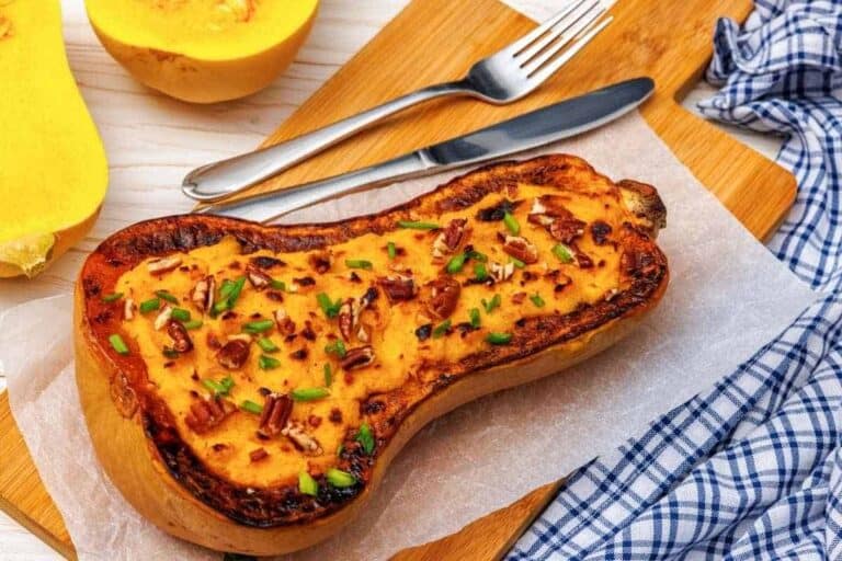 A baked stuffed butternut squash half topped with chopped herbs and pecans, placed on parchment paper with utensils and a blue plaid napkin nearby.
