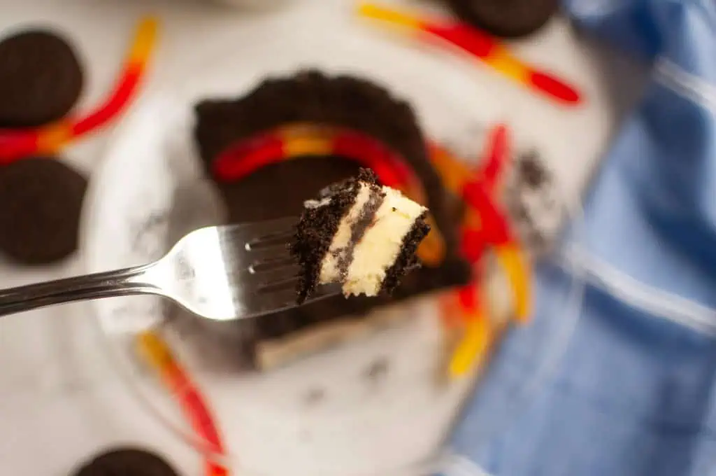 A fork holding a bite of Dirt cake, with gummy worms and crushed chocolate cookies on a plate in the background.