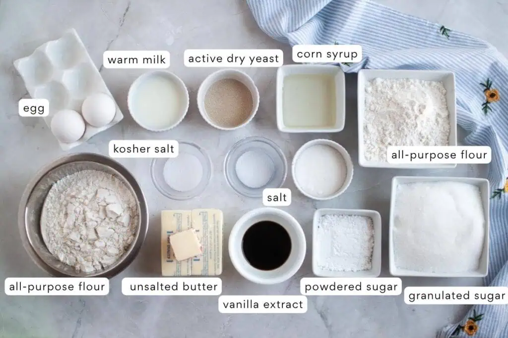 Assorted baking ingredients including eggs, milk, yeast, corn syrup, flour, salt, butter, vanilla, powdered and granulated sugar, displayed on a countertop.