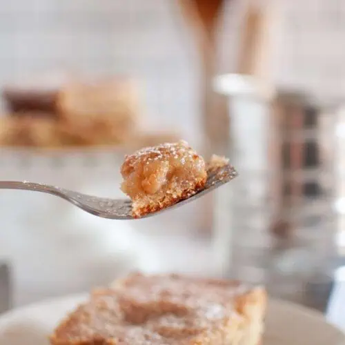 A fork holds a bite of powdered sugar-topped dessert bar above a plate with a full bar, with a blurred kitchen background.
