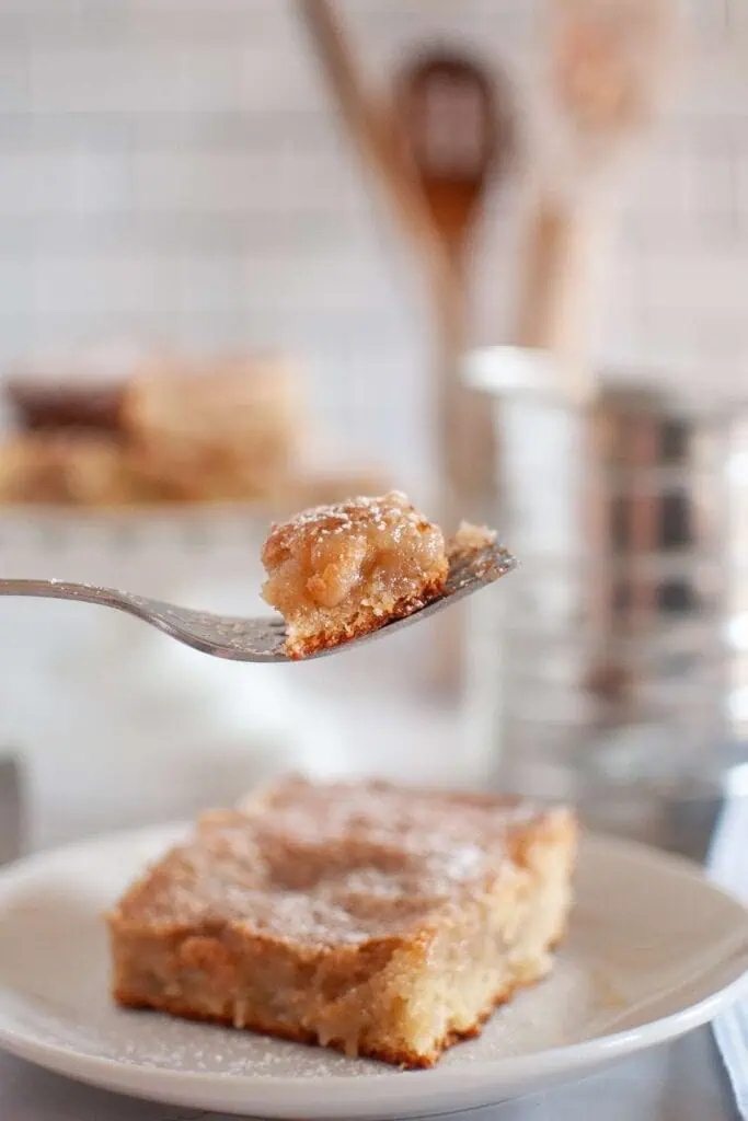 A fork holds a bite of powdered sugar-topped dessert bar above a plate with a full bar, with a blurred kitchen background.