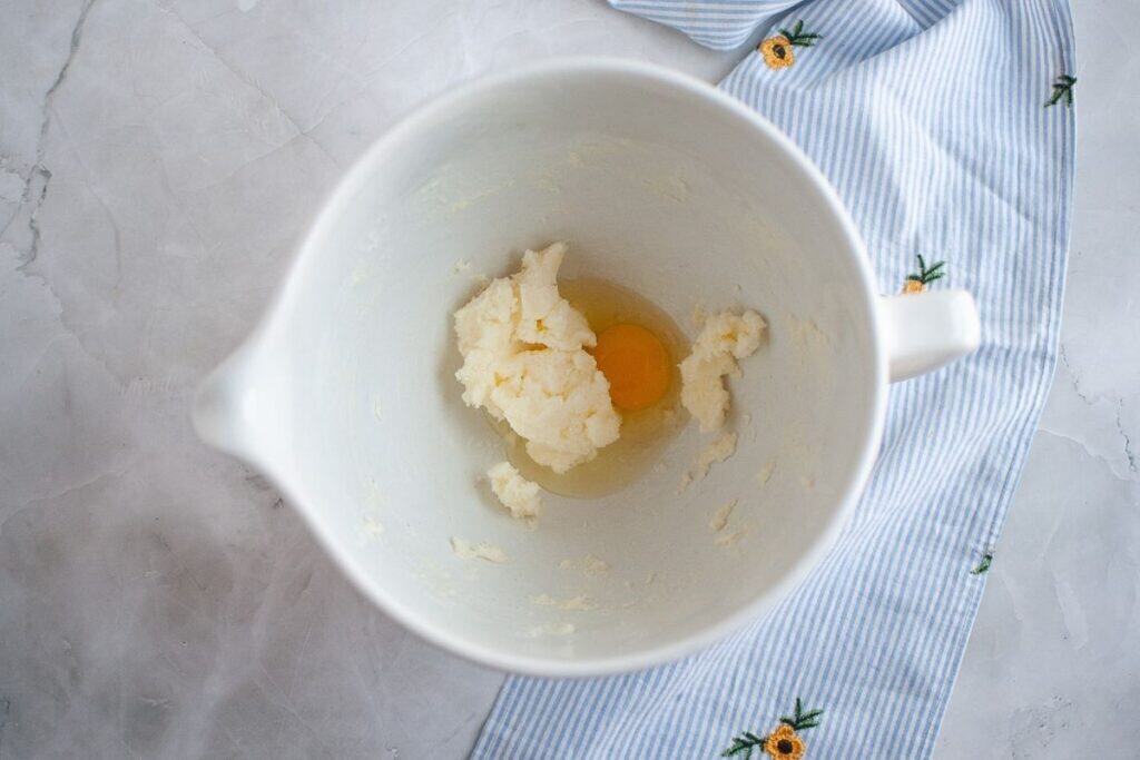 A mixing bowl containing creamed butter and sugar with a whole egg, placed on a marble surface next to a blue and white striped cloth with embroidered flowers.