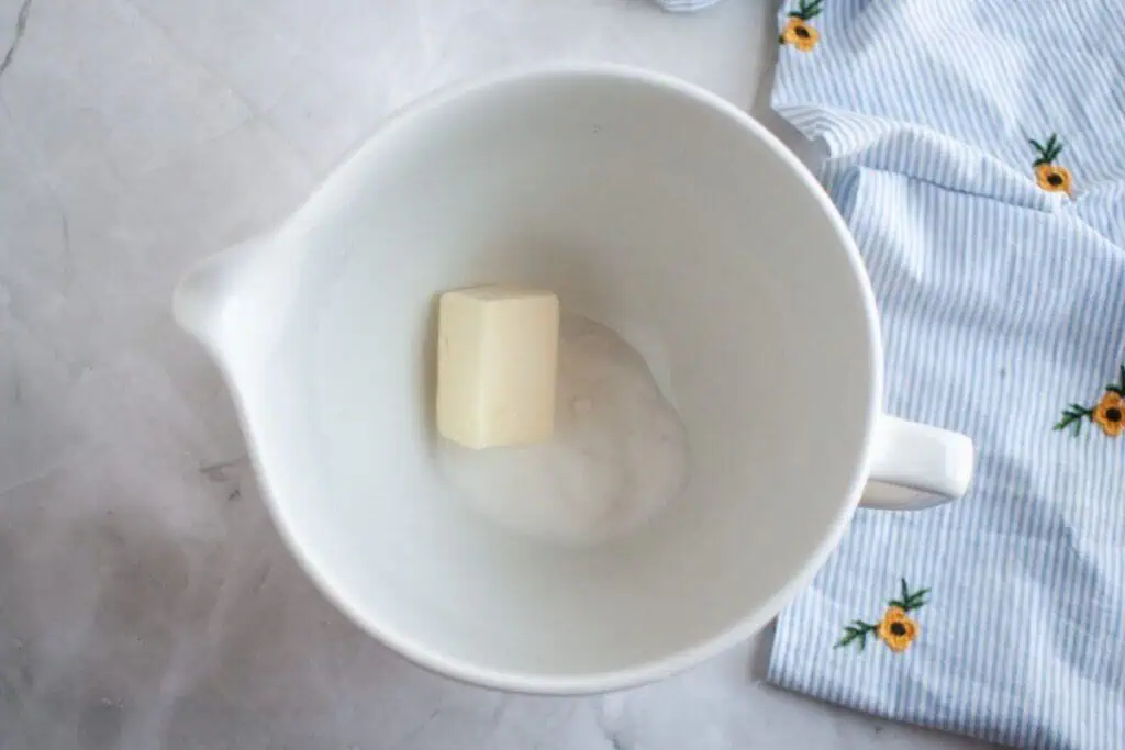 A white mixing bowl containing a piece of butter and some sugar sits on a marble surface next to a blue striped cloth with yellow flower designs.