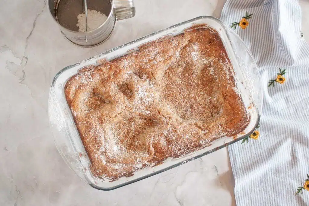 A glass baking dish filled with a golden-brown cobbler, lightly dusted with powdered sugar, sits on a marble surface next to a flour sifter and a striped cloth with floral embroidery.