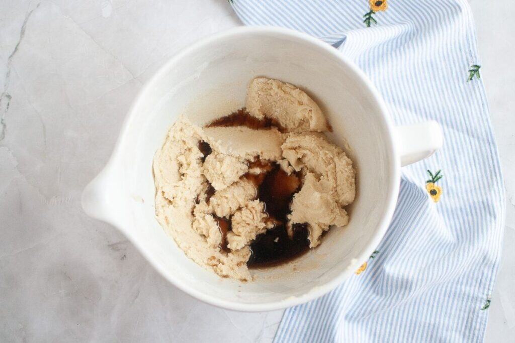 A white mixing bowl with partially mixed cookie dough and a pool of vanilla extract sits on a marble surface next to a blue and white striped cloth with flower patterns.