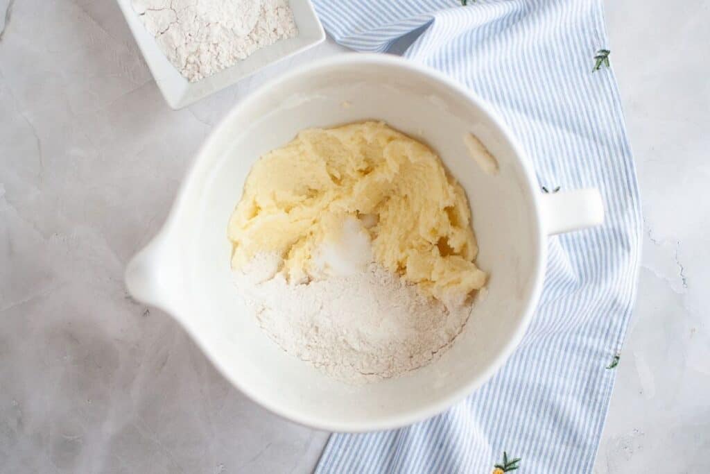 A mixing bowl with creamed butter and sugar and a portion of flour, set on a light surface with a blue striped cloth and a small dish of flour nearby.
