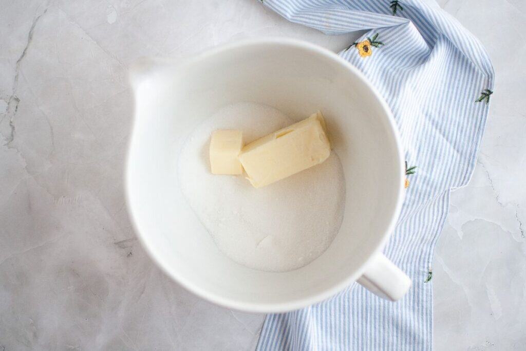 A white mixing bowl contains two sticks of butter and granulated sugar, placed on a light marble surface with a blue-striped cloth partially visible.