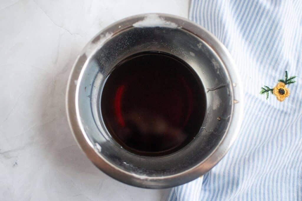 A metal bowl filled with a dark liquid sits on a white surface next to a blue and white striped cloth with a small embroidered sunflower.