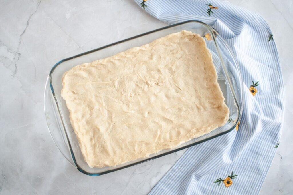 A rectangular glass baking dish with an even layer of unbaked dough, resting on a light marble surface next to a blue and white striped cloth with yellow flower embroidery.