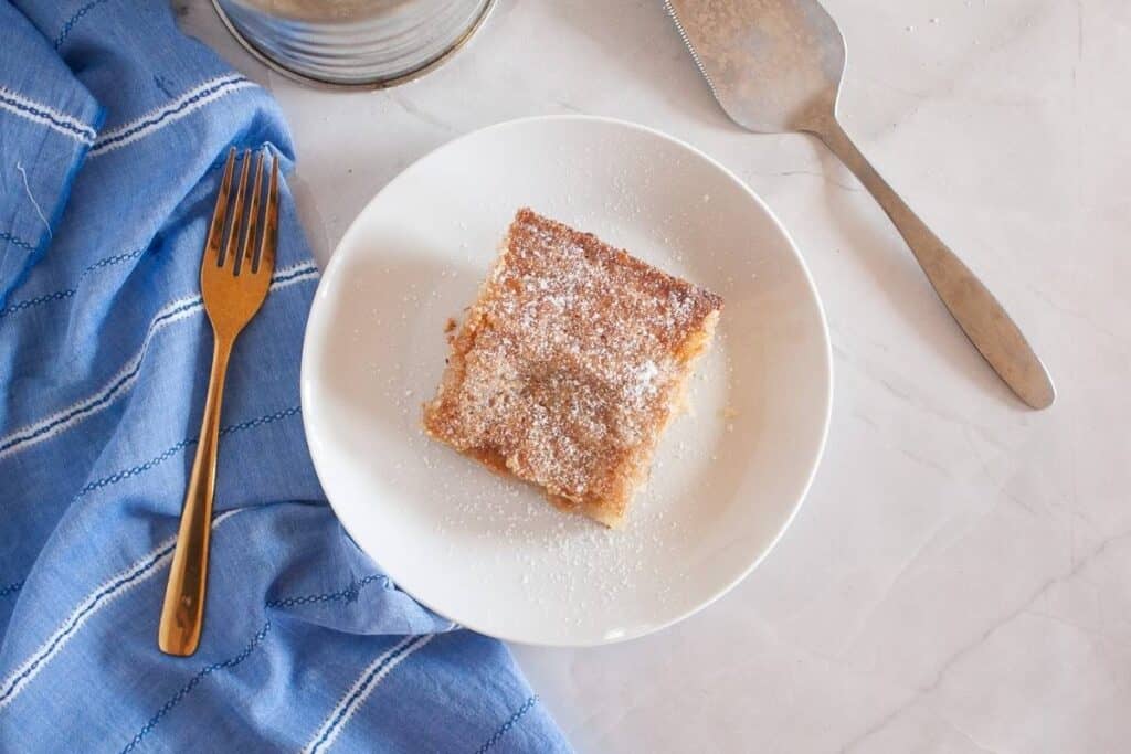 A square piece of cake with powdered sugar on a white plate, next to a gold fork, a blue cloth, and a metal cake server on a marble surface.
