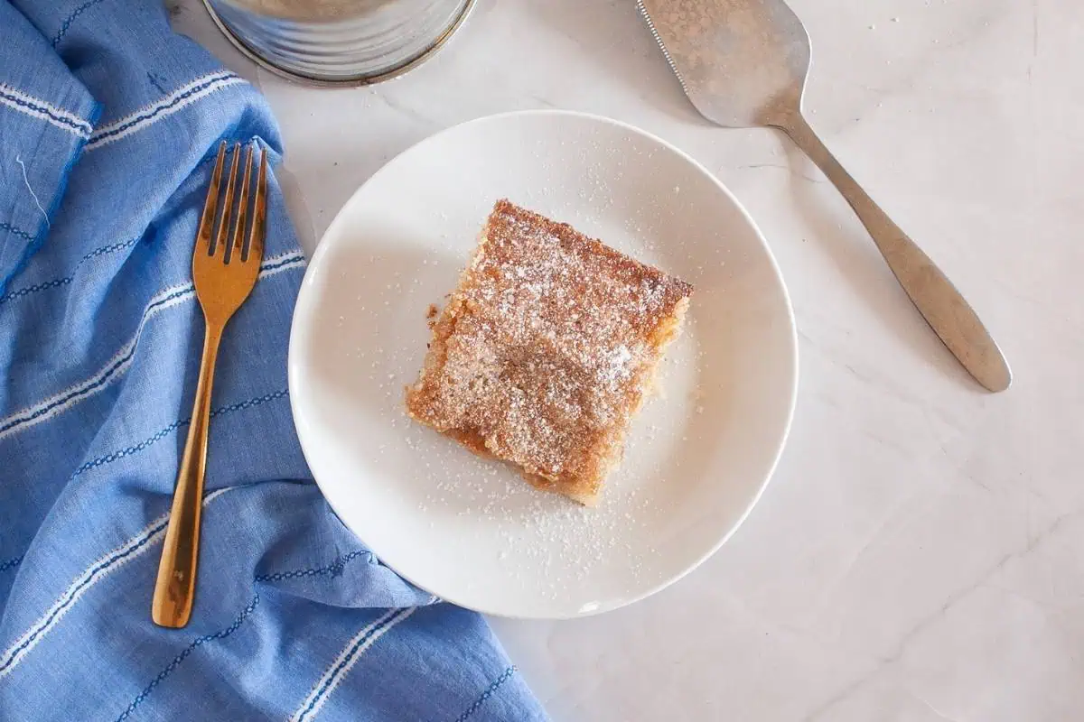 A square piece of Gooey Butter Cake. topped with powdered sugar on a white plate, next to a gold fork, a blue cloth, and a metal cake server on a marble surface.