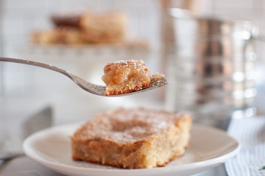 A fork holds a bite of a powdered sugar-topped dessert bar above a plate with a larger piece of the same dessert.