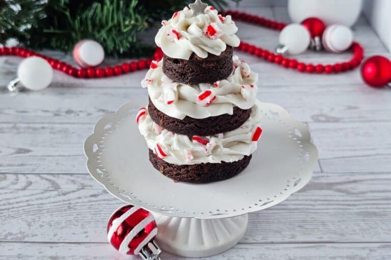A three-tiered chocolate brownie stack with white frosting and crushed peppermint, displayed on a white cake stand with Christmas ornaments in the background.