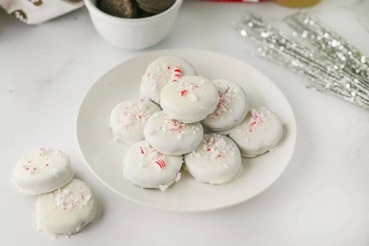 A white plate of Peppermint Crunch Oreos topped with crushed peppermint pieces sits on a white surface.