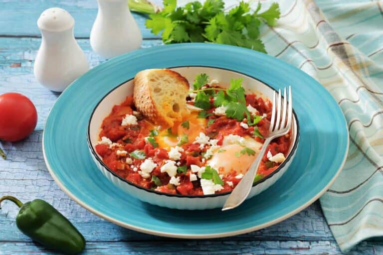 A bowl of shakshuka with poached eggs, tomato sauce, feta cheese, herbs, and a slice of toasted bread on a blue plate, with a fork and garnishes nearby.