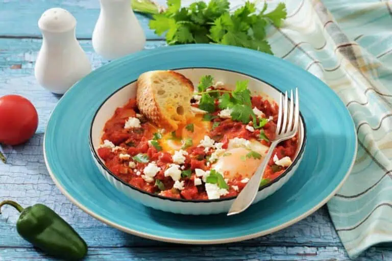 A bowl of shakshuka with poached eggs, tomato sauce, feta cheese, herbs, and a slice of toasted bread on a blue plate, with a fork and garnishes nearby.