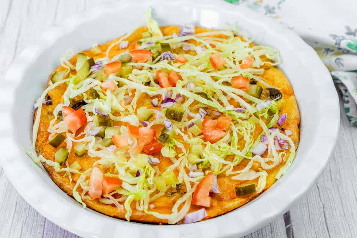A Cheeseburger Pie topped with shredded lettuce, diced tomatoes, pickles, and chopped red onions in a white pie dish on a light wooden surface.