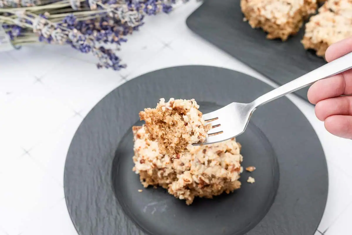 A fork holds a bite of Lazy Daisy Cake above a black plate with a cake square, with more cake pieces and dried lavender in the background.