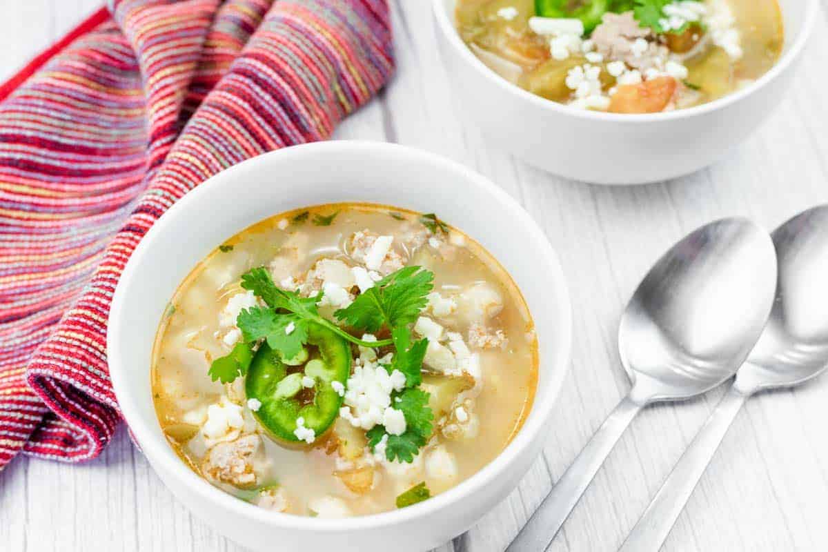 A bowl of Easy Green Pork Posole with white hominy, ground meat, sliced jalape&ntilde;os, cheese, and cilantro, next to a striped cloth napkin and two metal spoons on a white surface.