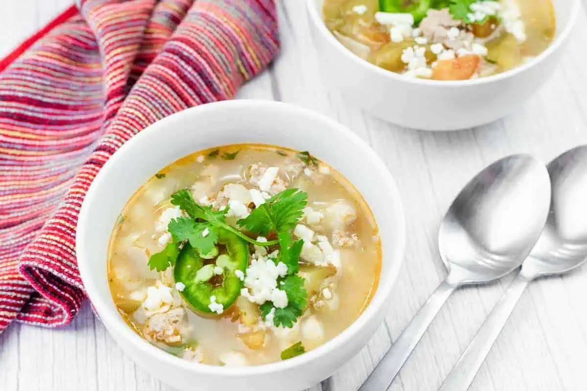 A bowl of Easy Green Pork Posole with white hominy, ground meat, sliced jalapeños, cheese, and cilantro, next to a striped cloth napkin and two metal spoons on a white surface.