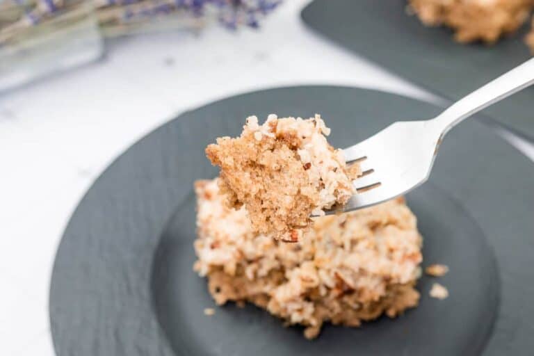 A fork holds a bite of crumbly cake with icing above a plate containing the remaining piece of cake.