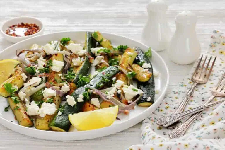 A plate of roasted zucchini with red onions, feta cheese, parsley, and lemon wedges, served on a white table with salt and pepper shakers, a napkin, and forks.