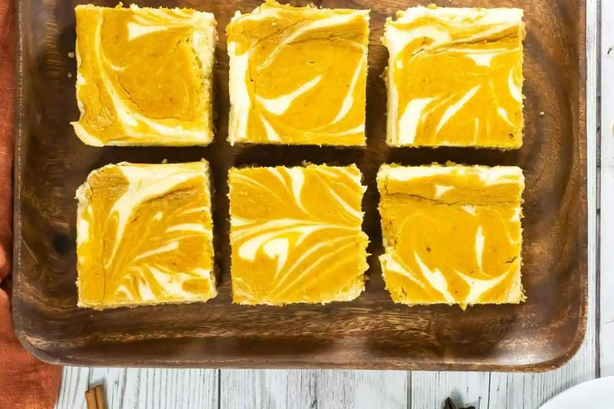 Six square pumpkin cheesecake bars are arranged on a wooden tray, displaying marbled orange and white patterns on top.