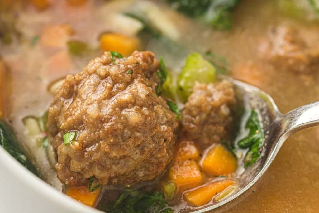 A close-up of a spoon holding a meatball above a bowl of soup with chopped carrots, greens, and broth.