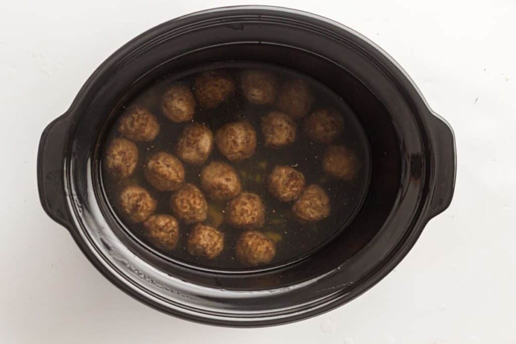 A black slow cooker filled with meatballs submerged in a clear broth or liquid.