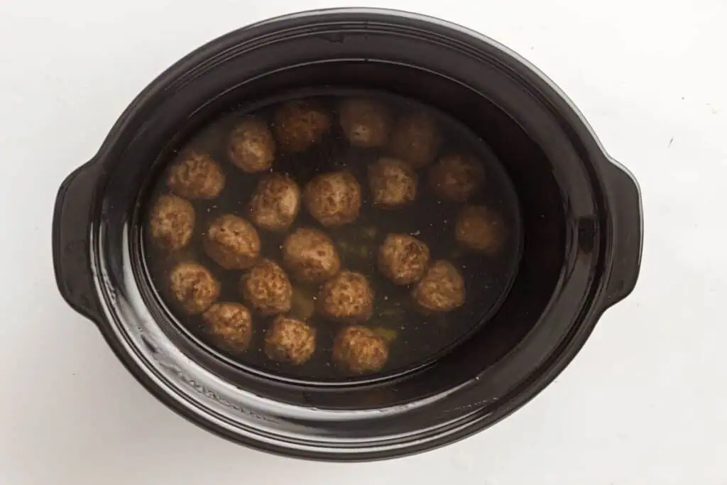 A black slow cooker filled with meatballs submerged in a clear broth or liquid.