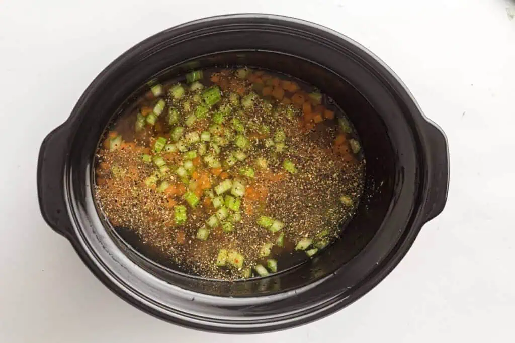 A slow cooker filled with chopped carrots, celery, broth, and seasonings, ready to be cooked.