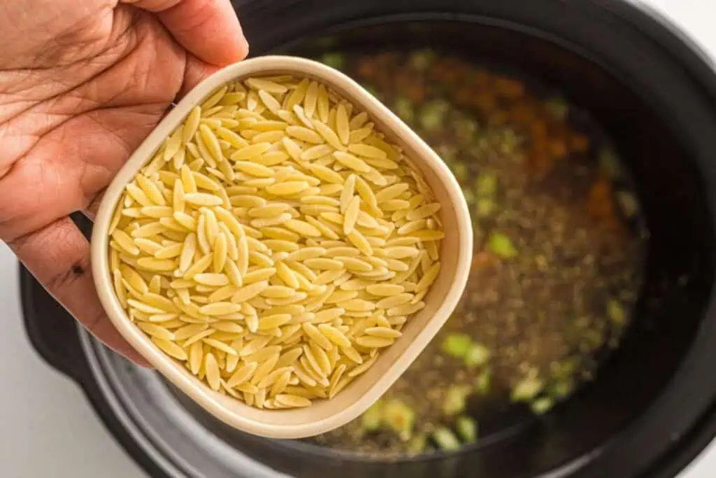 A hand holds a bowl of uncooked orzo pasta over a slow cooker filled with broth and chopped vegetables.