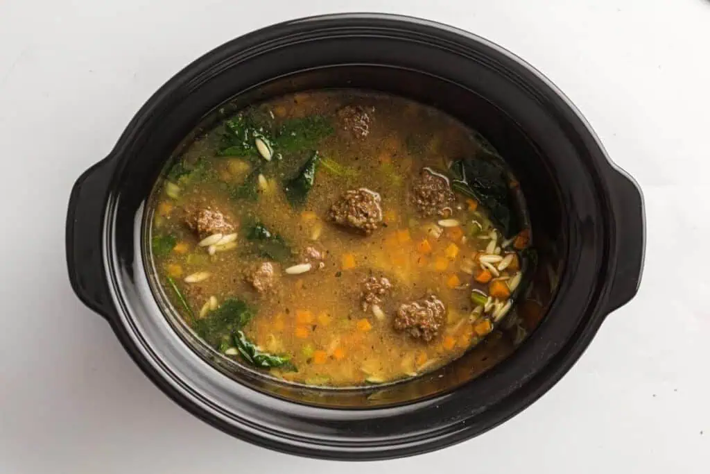 A black slow cooker filled with soup containing meatballs, spinach, diced carrots, orzo pasta, and broth on a white background.