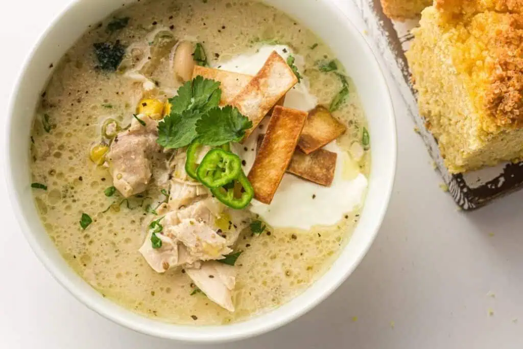 A bowl of creamy chicken chili topped with sliced jalapeños, tortilla strips, sour cream, and cilantro, next to a plate of cornbread.