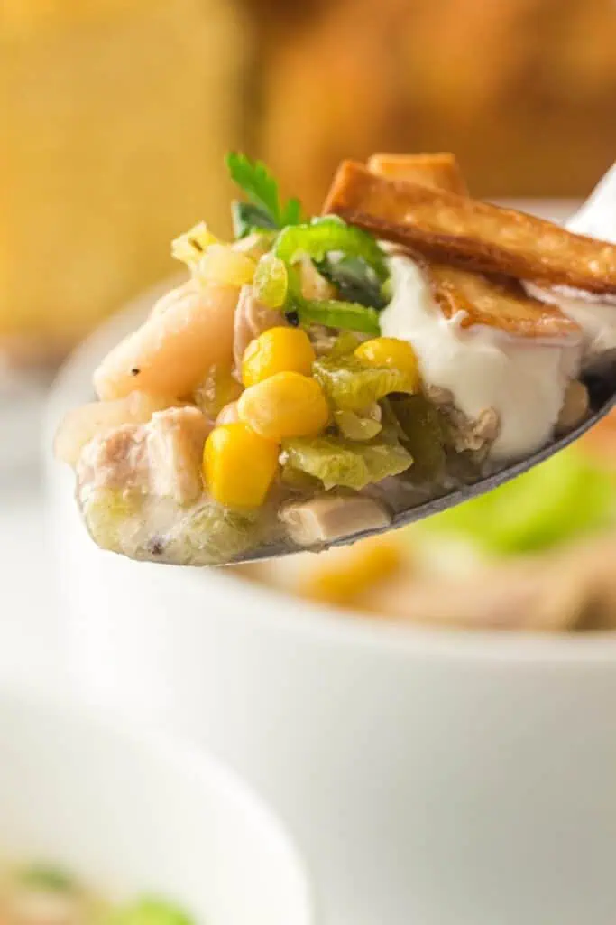 A close-up of a spoonful of chicken chili with corn, green chilies, beans, sour cream, parsley, and tortilla strips.