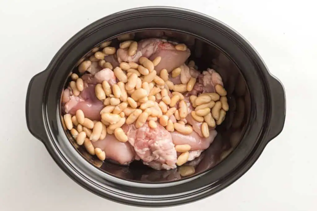 Raw chicken pieces and white beans placed inside a black slow cooker, viewed from above on a white background.
