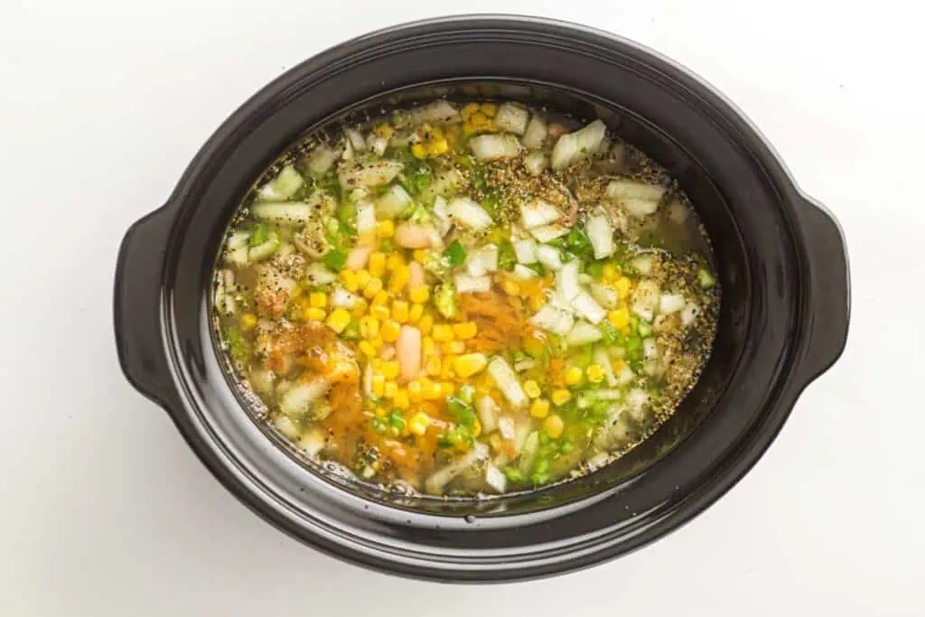 A slow cooker filled with chopped onions, corn, beans, carrots, seasonings, and broth, ready to be cooked.