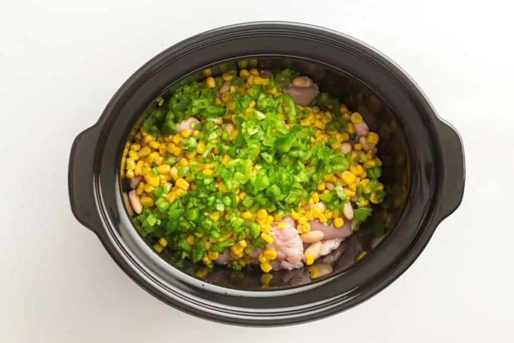 A slow cooker filled with raw ingredients including corn, white beans, chopped green peppers, and pieces of chicken, viewed from above.