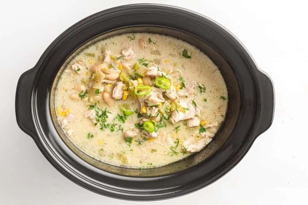A black slow cooker filled with creamy white chicken chili, garnished with chopped herbs and sliced green chilies.