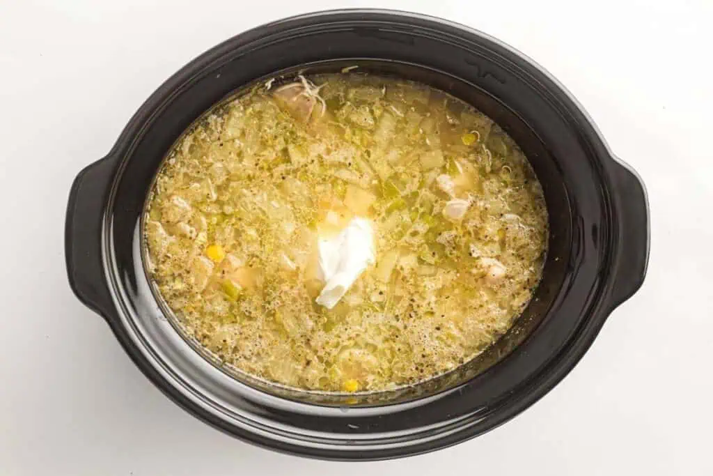 A slow cooker filled with a chunky soup or stew, topped with a dollop of sour cream, photographed from above on a white background.