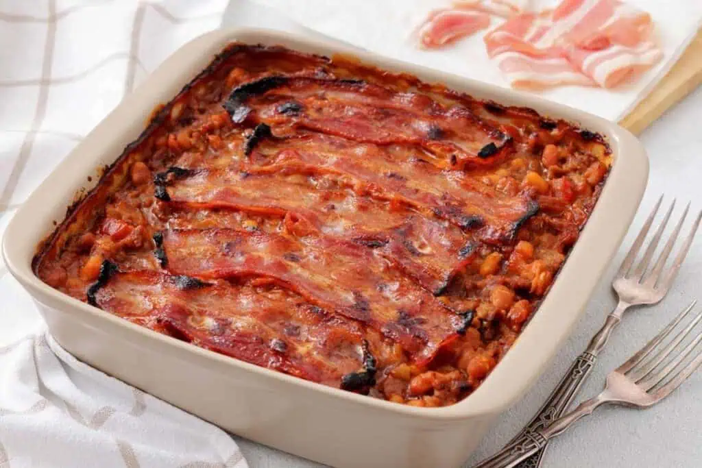 A casserole dish filled with baked beans and topped with strips of cooked bacon. Two forks and slices of raw bacon are placed nearby.