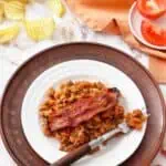 A white plate with baked beans and a strip of bacon sits on a wooden charger. Nearby are potato chips, sliced tomatoes, and an orange napkin.
