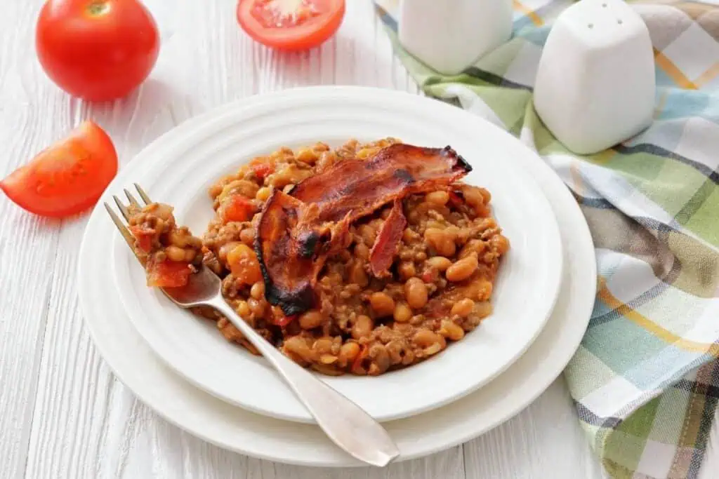 A white plate with baked beans topped with a slice of bacon, next to a fork, tomato wedges, salt and pepper shakers, and a plaid napkin on a wooden table.